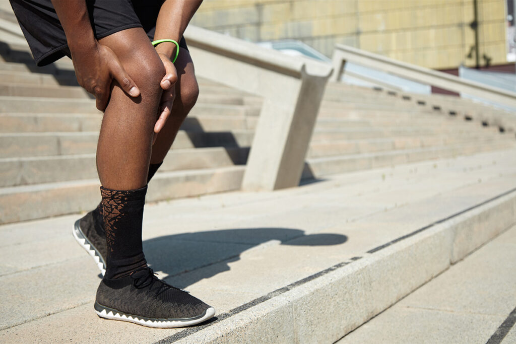 Foto focada nas pernas e joelhos de uma pessoa negra. Ela está com as mãos sobre o joelho, usa bermuda, tênis e meia.